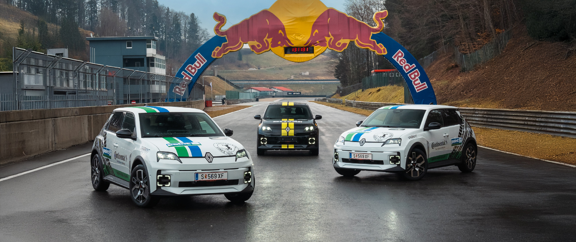 Drei sportliche Renault-Fahrzeuge stehen auf einer Rennstrecke unter einem Red Bull-Bogen. Zwei weiße Fahrzeuge mit grünen und blauen Akzenten tragen Logos von Sponsoren wie Continental und Point S, während das mittlere schwarze Auto gelbe Rennstreifen hat.
